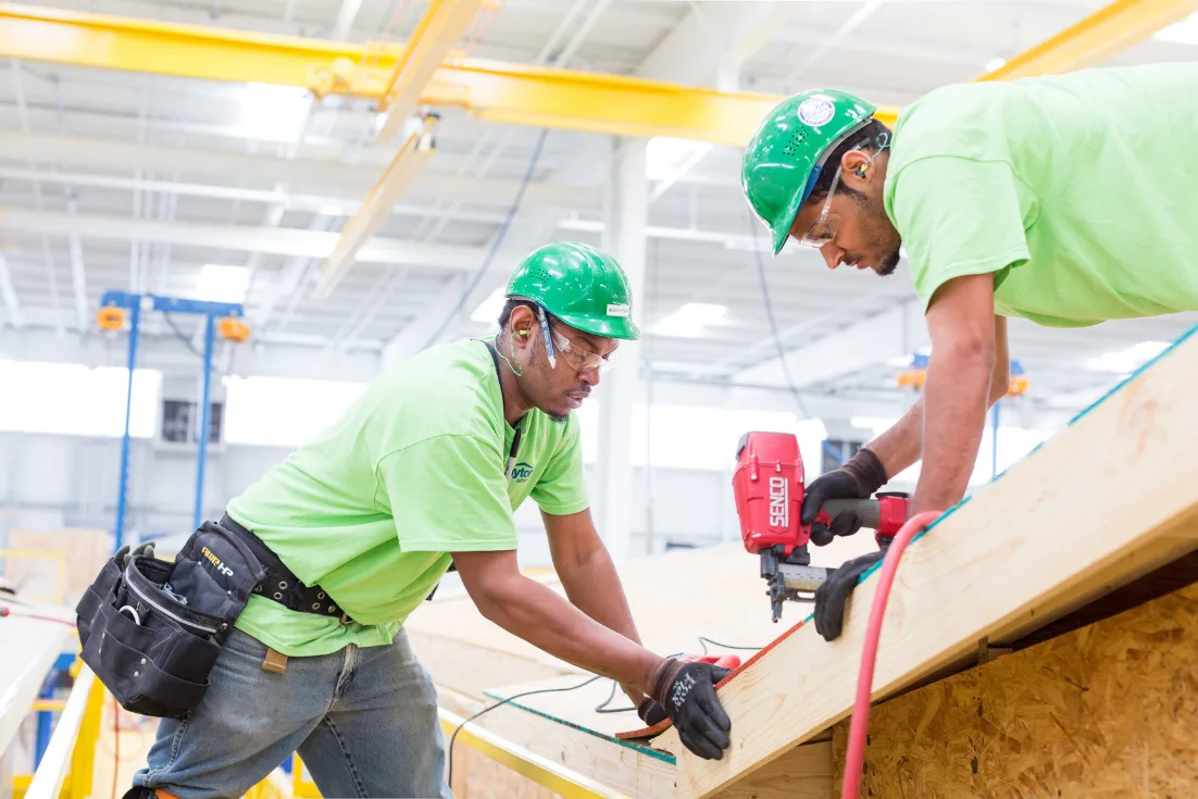 Two facility workers in bright green shirts, safety glasses, and and dark green construction hats hold a home frame while one drills into it.