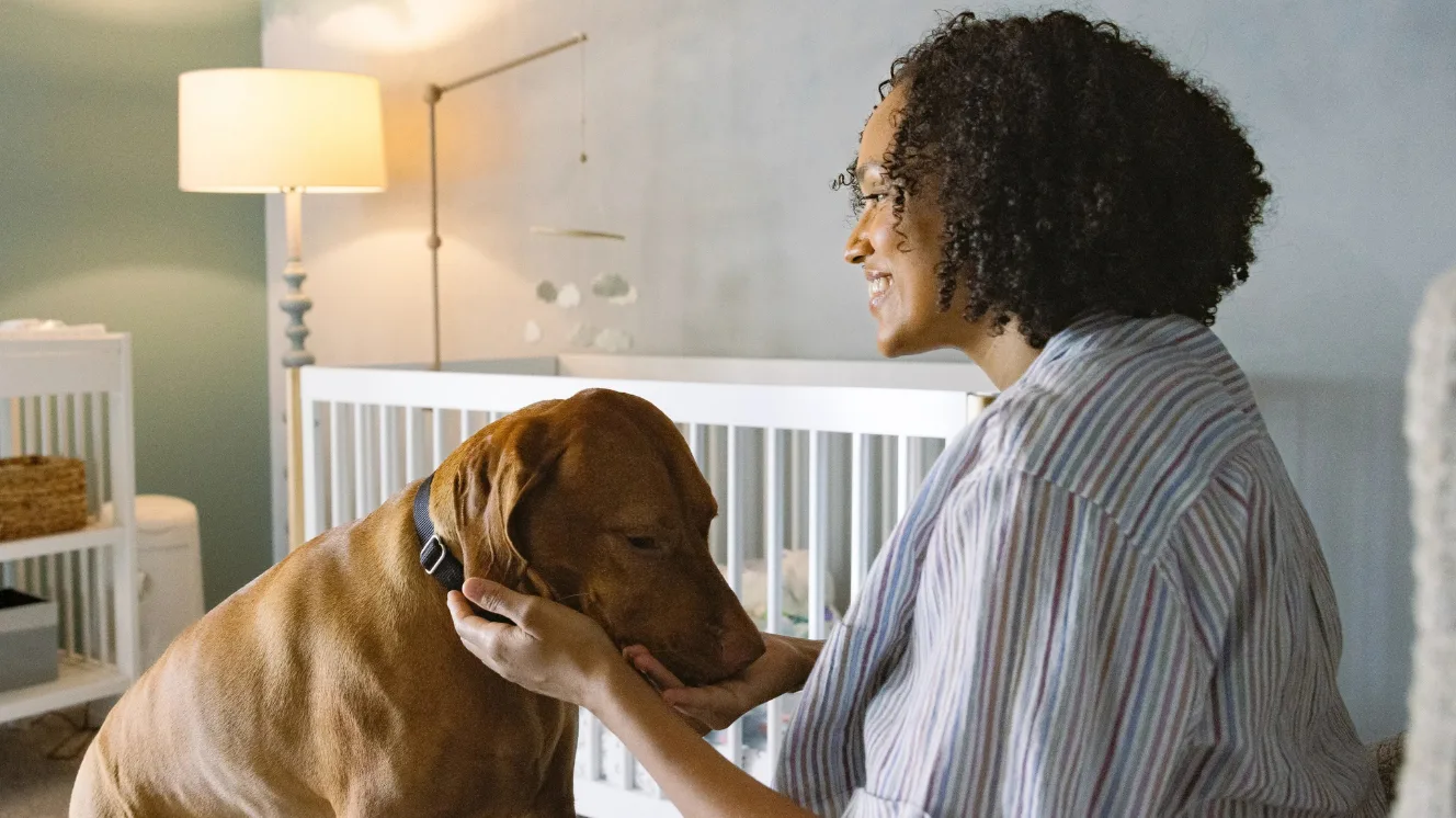 A woman on the right sits and smiles while petting her tan dog in a nursery room.