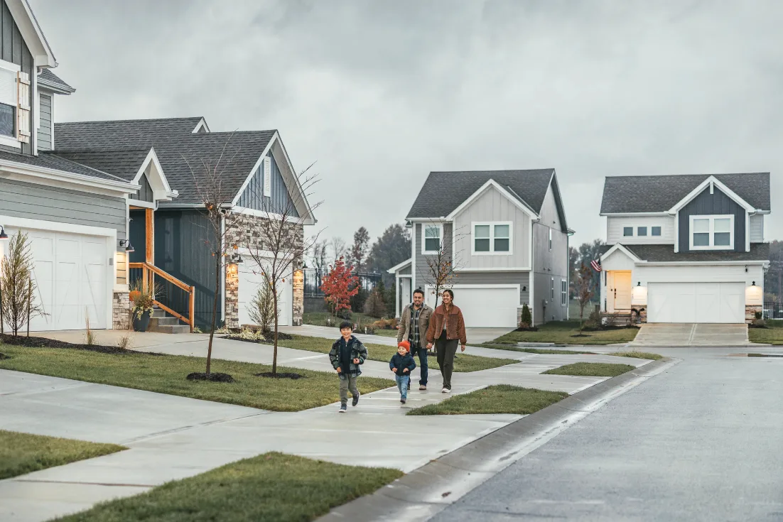 A family of four in jackets walks down the sidewalk of neighborhood with new two-story site-built homes in shades of gray and blue behind them on an overcast day.