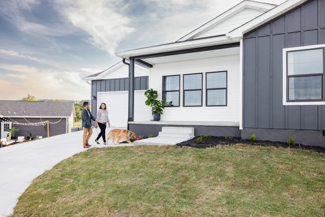 A couple walks their dog on a leash up the walkway to their white and gray home's front porch.