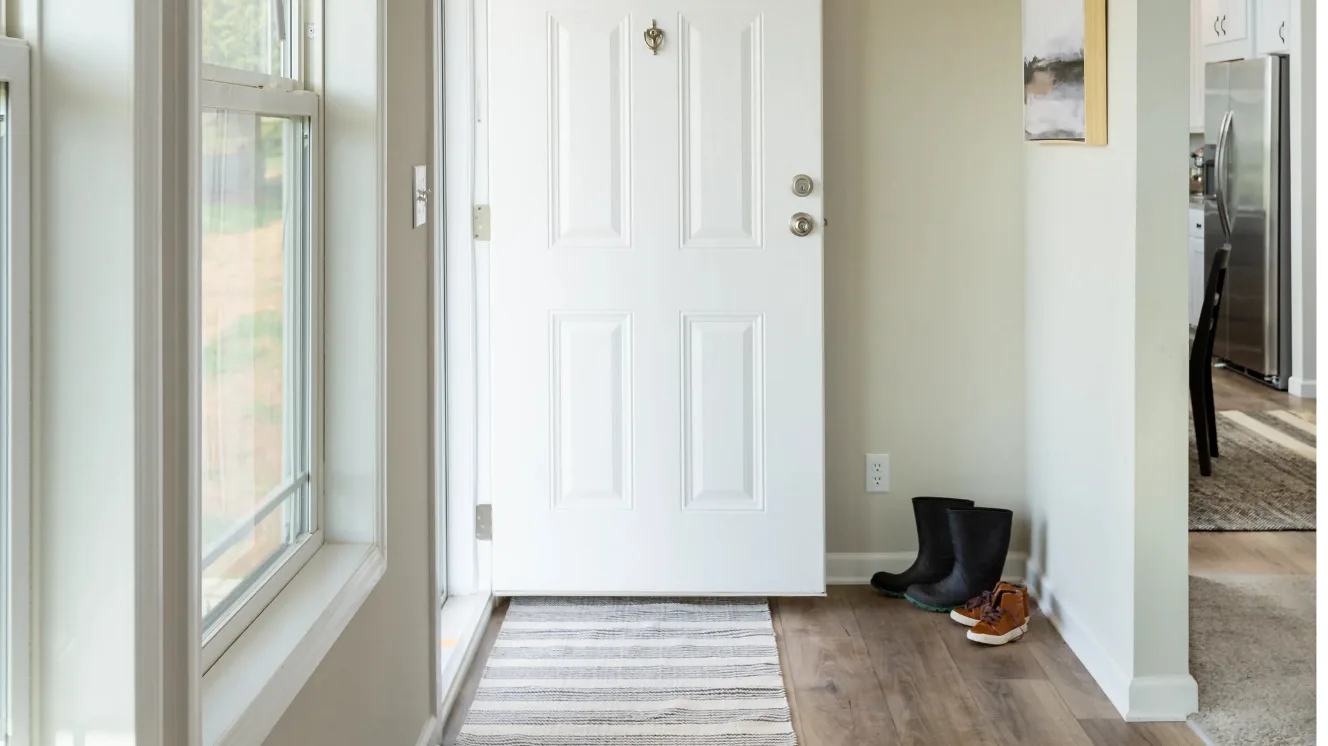 Inside the foyer of a home, the front door is open, two pairs of shoes are beside it, and the kitchen entry is seen in the background.