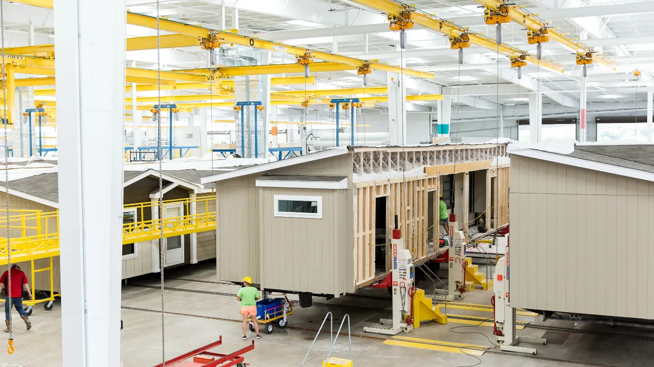 Inside of a manufactured home building facility with a white ceiling and concrete floor, with people working on three different home sections.
