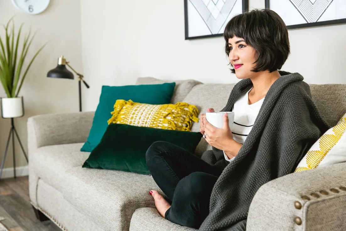 A woman sits with her legs crisscrossed on a beige couch beside colorful pillows with a dark blanket pulled over her shoulders and a mug in her hand.