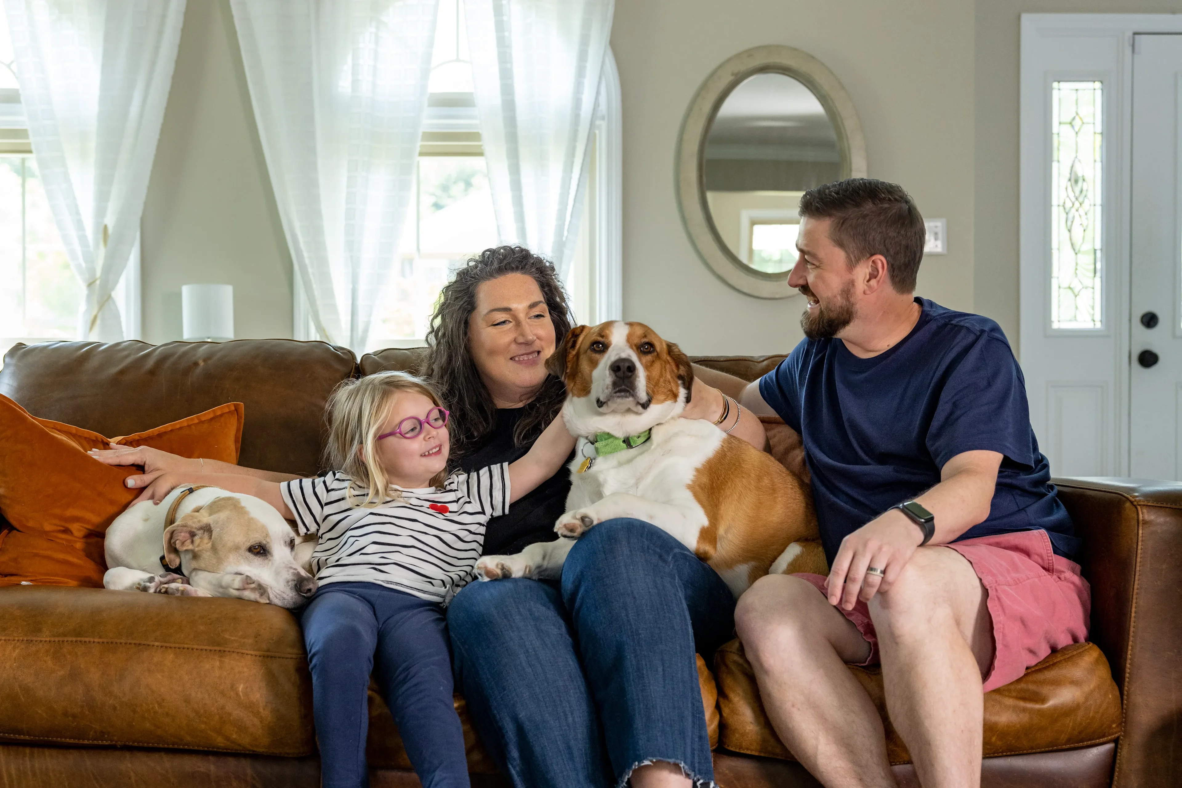Woman, man and young girl sit on a leather couch with two dogs, with windows in the background.