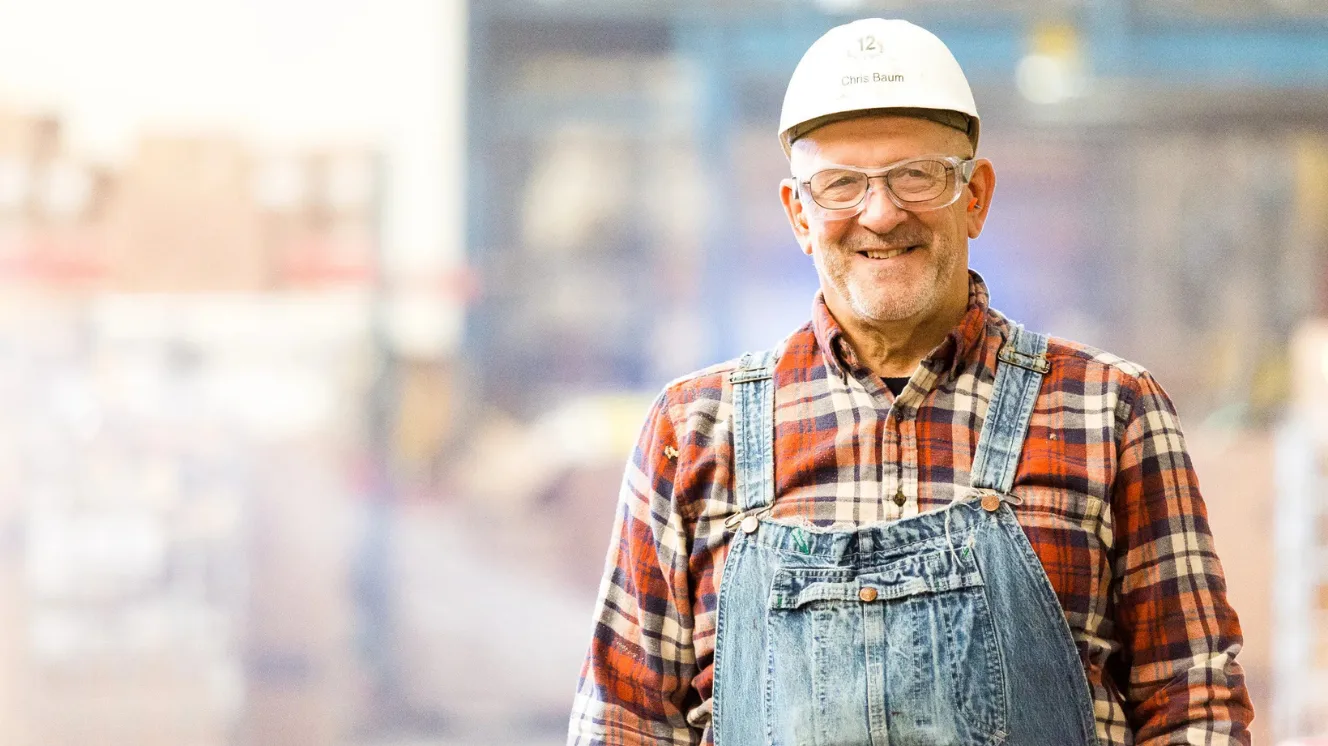 A man in a plaid flannel button up and overalls smiles while wearing safety glasses and a white construction safety hat.