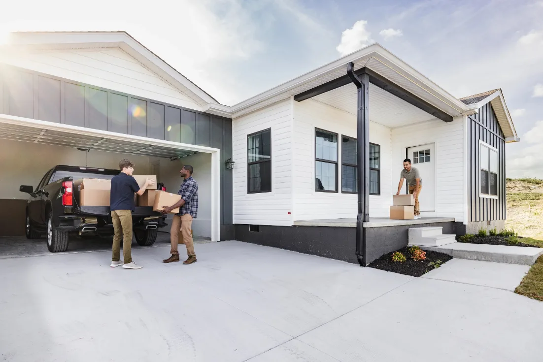 Two men unload moving boxes from a black truck parked in an open garage, while another man picks up boxes off the porch of a new home in front of the door.