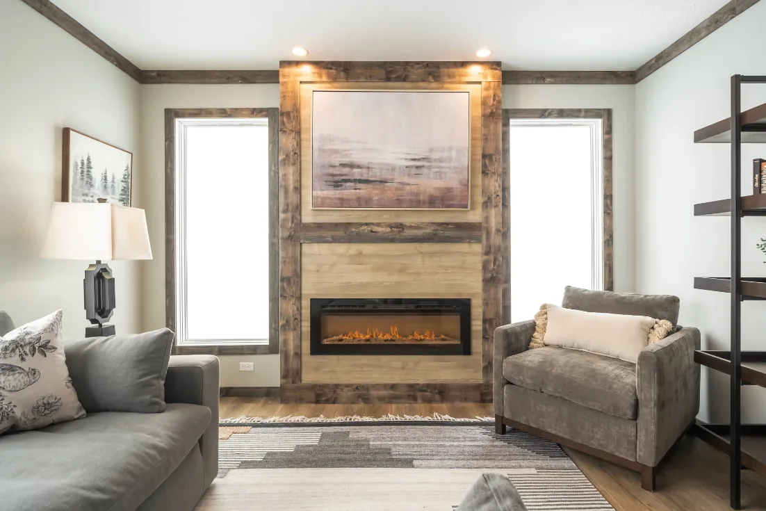 An abstract painting hangs over an electric fireplace between two tall windows and in the center of a living room furnished with a gray couch and chair.