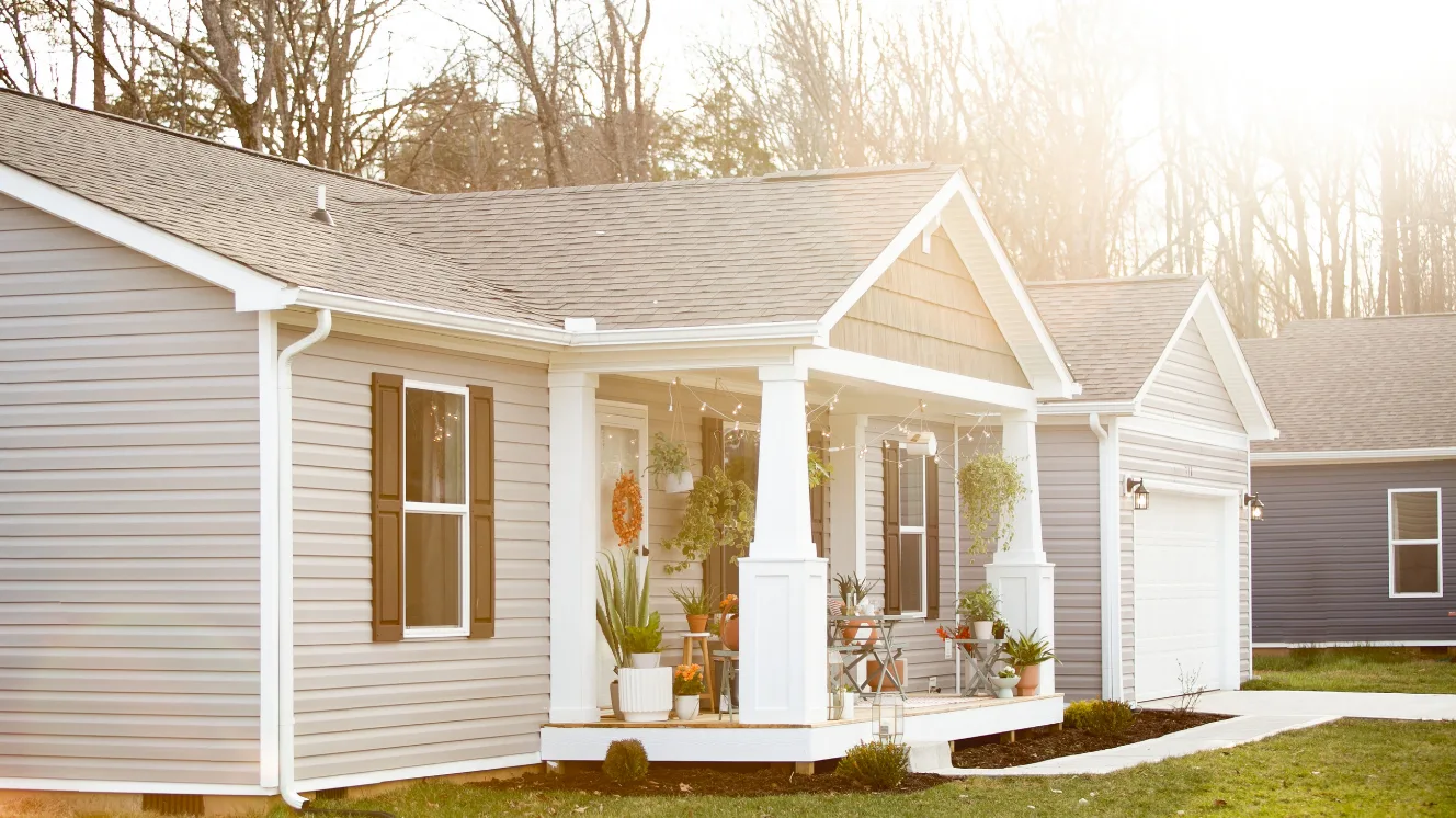 The exterior of modern manufactured home with gray siding, a front porch with thick white columns and potted plants decorating it, and an attached garage.