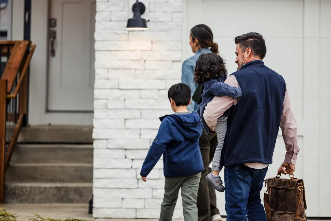 A family of four, the dad carrying the youngest boy, walk to their white brick home's front door.