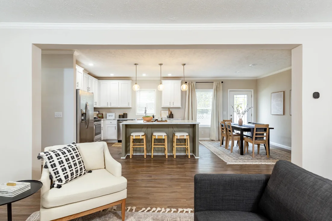 A view of a kitchen from the living room of a Clayton CrossMod home with cream walls, white cabinets, a large sage green kitchen island with seating in the center and a dining table set on the right.