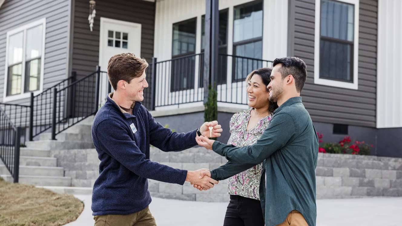 A home consultant in a blue sweater shakes hands and hands over a key to a couple in front of a gray and white home.