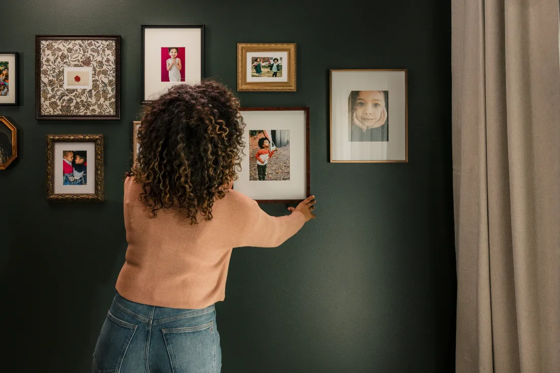 The back of a woman in a tan sweater is shown as she hangs up a photo of a child on a gallery wall of family photos.