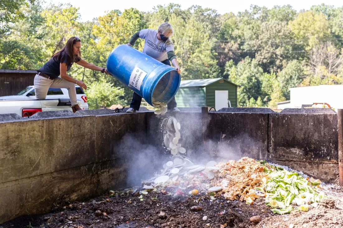 Two women dump gypsum from a blue recycling bin into a large outdoor compost container.