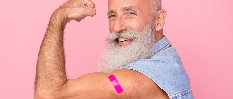 Middle aged man flexing muscles with flu jab plaster on one arm against pink background.