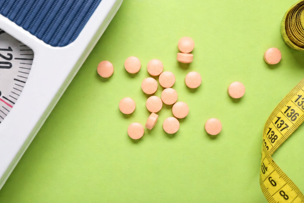 Pink tablets scattered on a green surface beside a white weighing scale and a yellow measuring tape.