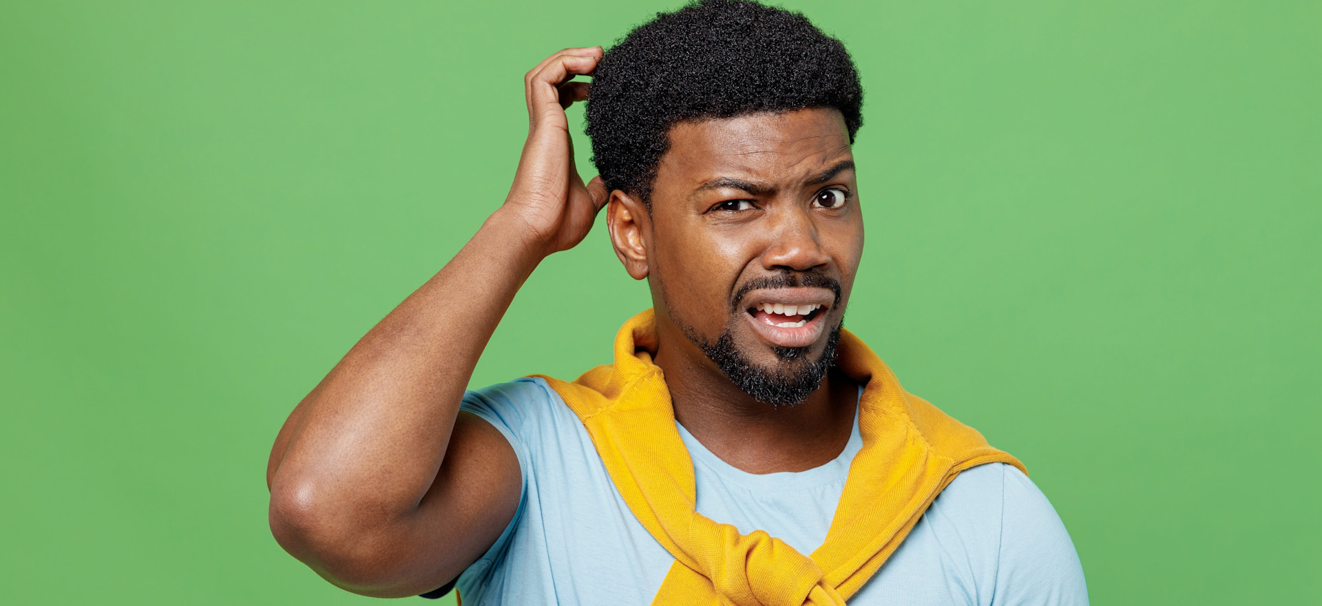 Person with curly hair scratching head with confused expression, wearing yellow hoodie against bright green background.