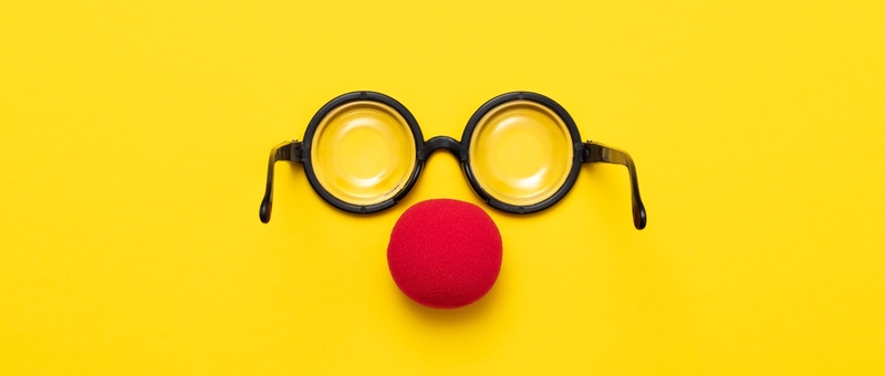 Red clown nose beneath round, black-rimmed comedy glasses against a yellow background.