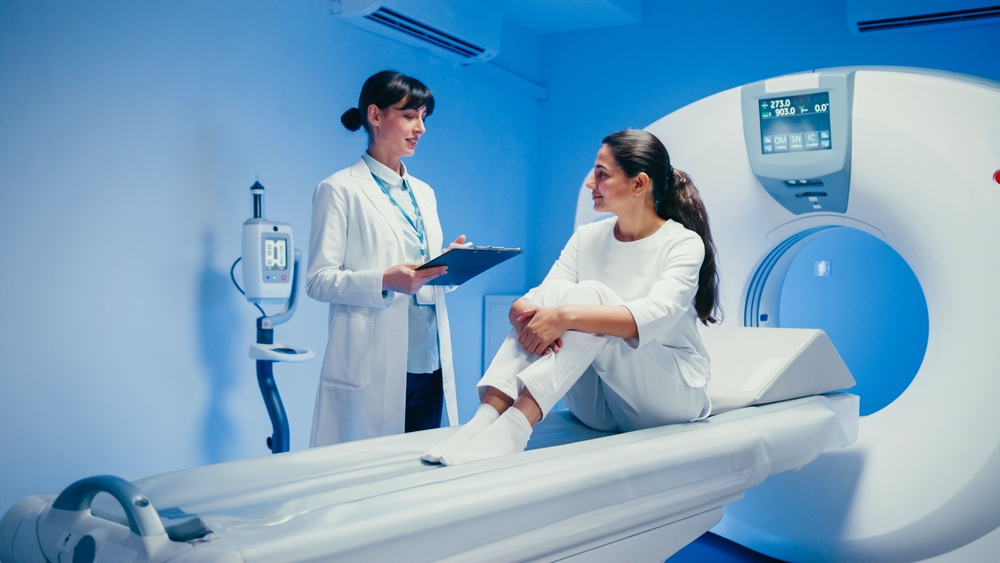 Relaxed woman sitting on MRI table nodding head while listening to female doctor explaining. Holding clipboard. Friendly interaction happening in clinical setting during preparation for radiology scan