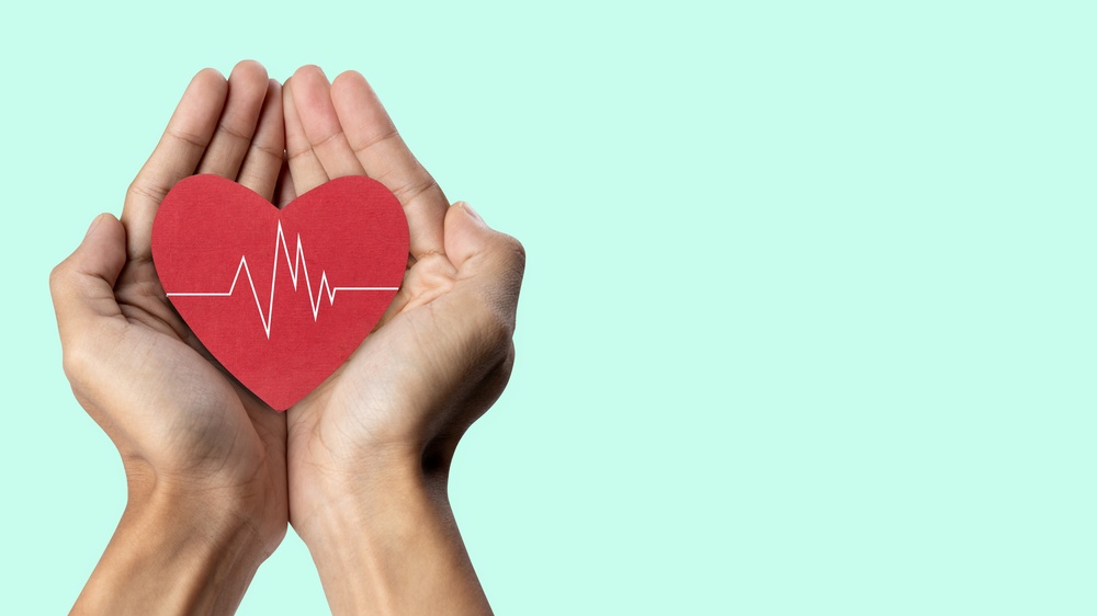 A pair of hands holds a red heart shape, symbolizing love, health, and compassion. The heart displays a heartbeat line, representing life and healthcare