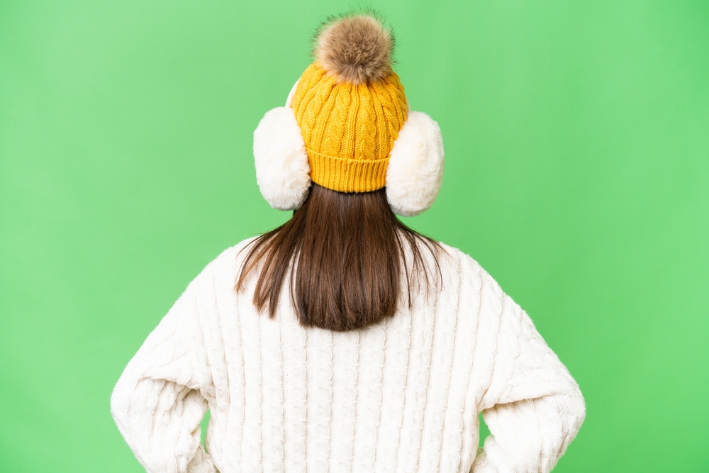 Rear view of brown-haired girl wearing white winter earmuffs, bobble hat, and knitted jumper against green background.