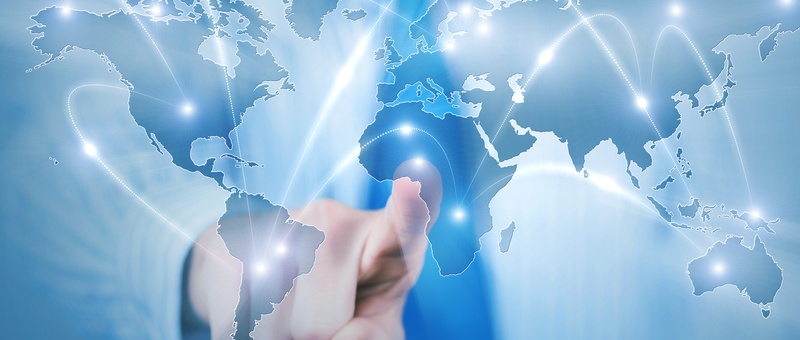 Hand touching a digital world map with glowing connection lines linking continents on a blue background.