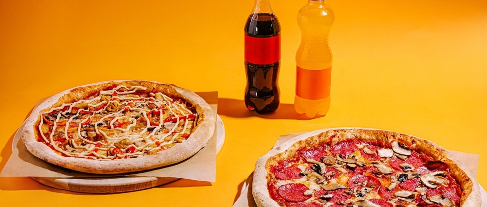 Two mouthwatering pizzas are displayed with a colourful backdrop. One pizza features pepperoni and mushrooms, while the other has a creamy topping. Bottles of drinks complement the meal.