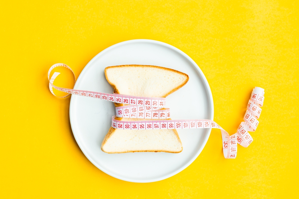 A slice of white bread wrapped tightly with a measuring tape on a white plate, set against a bright yellow background.