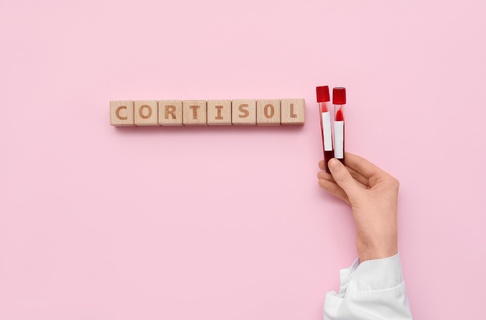 Wooden blocks spelling "CORTISOL" with a hand in a white coat holding two blood test tubes against a pink background.