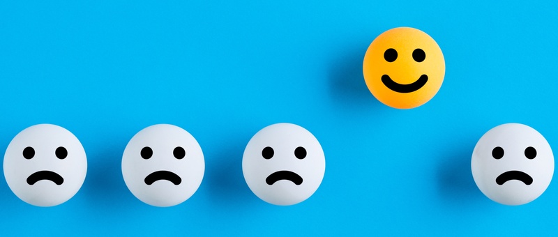 Four sad white circular faces in a line with one smiling yellow circular face elevated above the rest against a bold blue background.