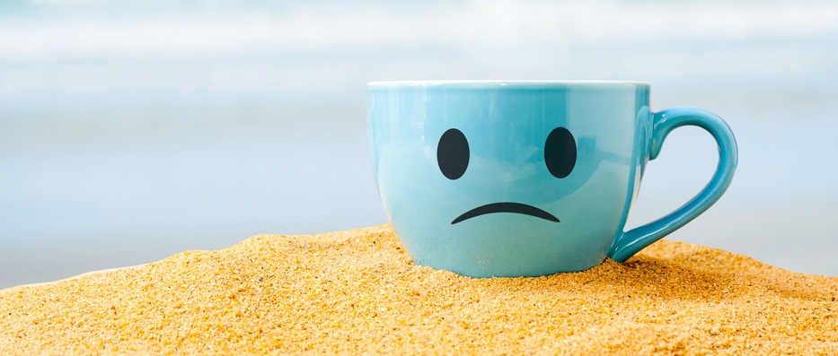 blue cup of coffee with unhappy face on white sand beach over blue sky and sea on day noon light background
