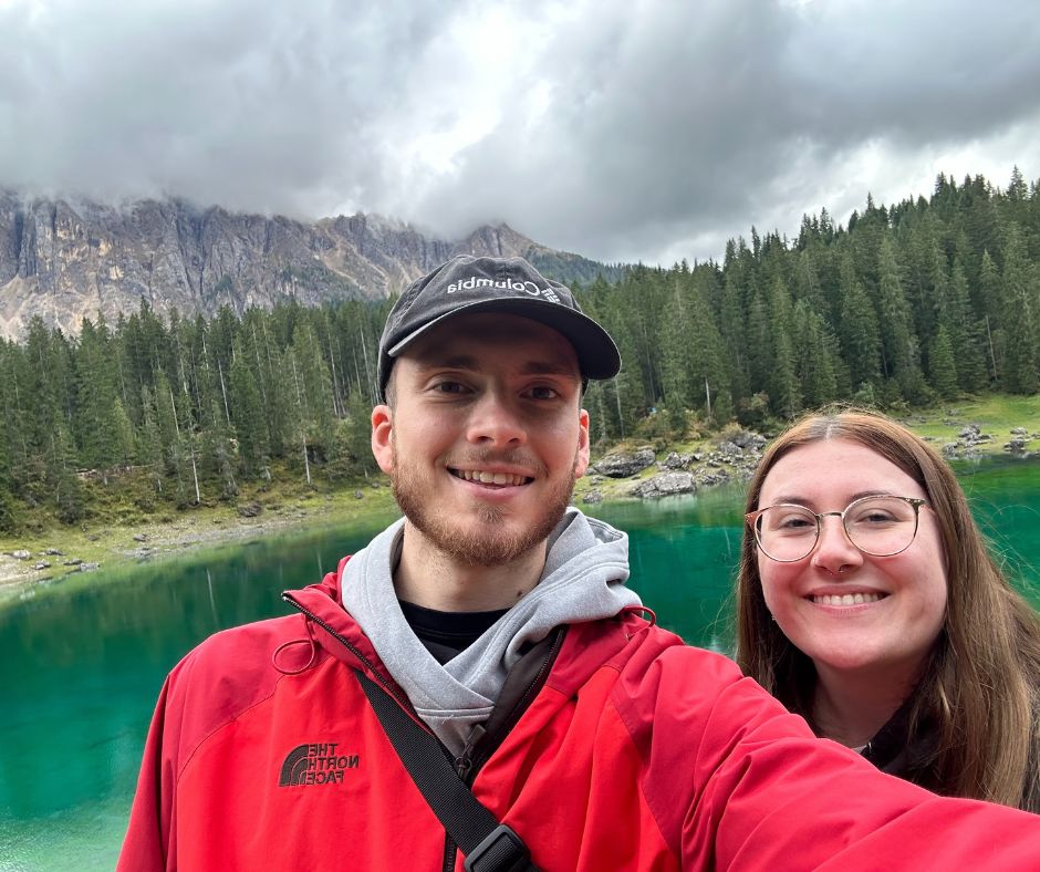 Young couple posing for a selfie against a tree-lined lake beneath mountain range.