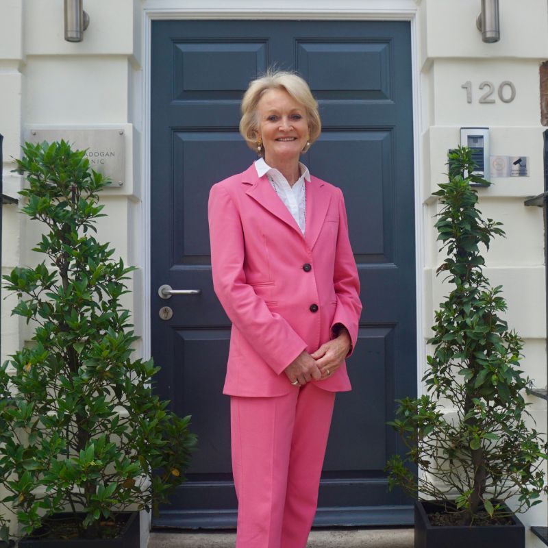 Dr Susan Mayou, Consultant Dermatologist at Cadogan Clinic wearing pink trouser suit, standing outside clinic doorway.