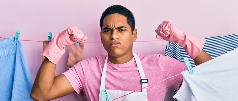 Young hispanic man wearing cleaner apron holding clothes on clothesline showing arm muscles and proudly smiling.