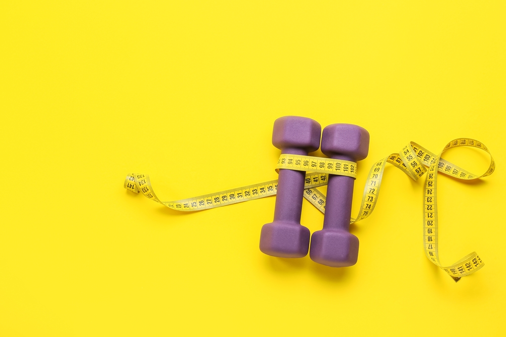 A purple dumbbell wrapped with measuring tape, standing upright against a bright yellow background.