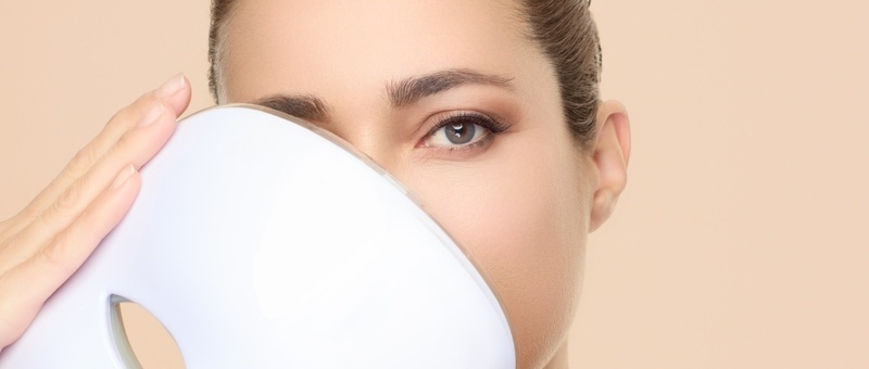 Woman holding LED mask close to her face against a pale peach background.