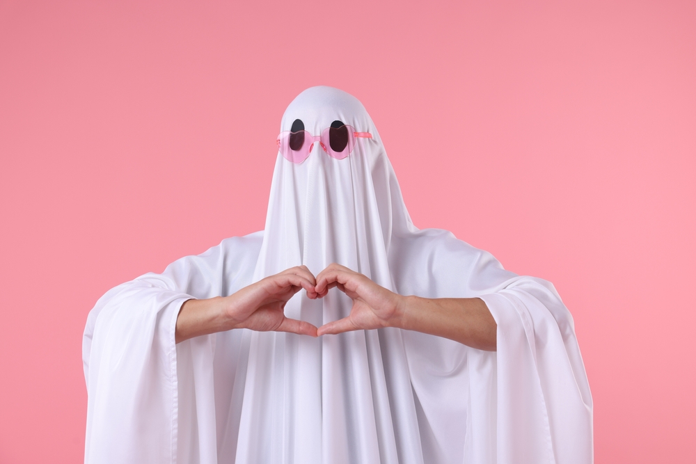 Man covered with white ghost sheet in pink heart-shaped sunglasses making heart hands gesture against pink background.