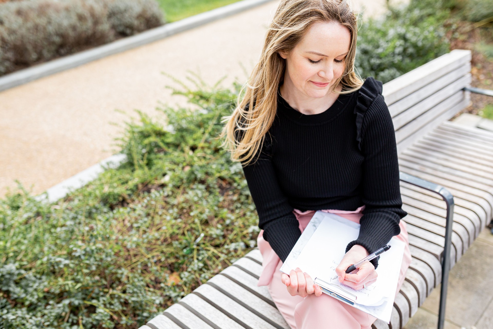 Female accredited psychotherapist Laura Greenwood of Laura Greenwood Therapy sitting on a bench in nature with notepad. 