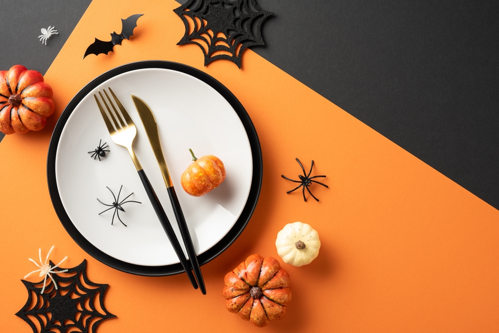 Halloween table setting, view from above. Flat lay black empty plate, cutlery, spooky decorations, bats, spiders, pumpkins on orange and black background.