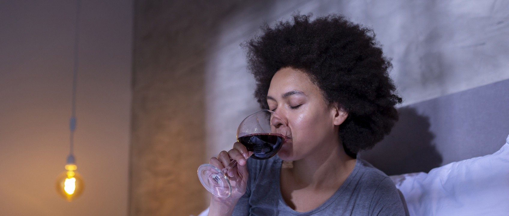 Person with curly hair sipping red wine from a glass in a softly lit room with a warm pendant light visible.