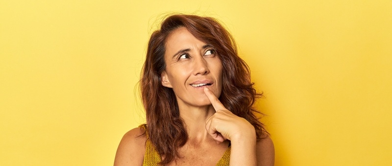 Middle-aged woman on a yellow backdrop looking sideways with doubtful and skeptical expression.