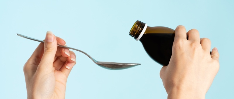Pair of hands holding cough syrup and spoon against blue background.