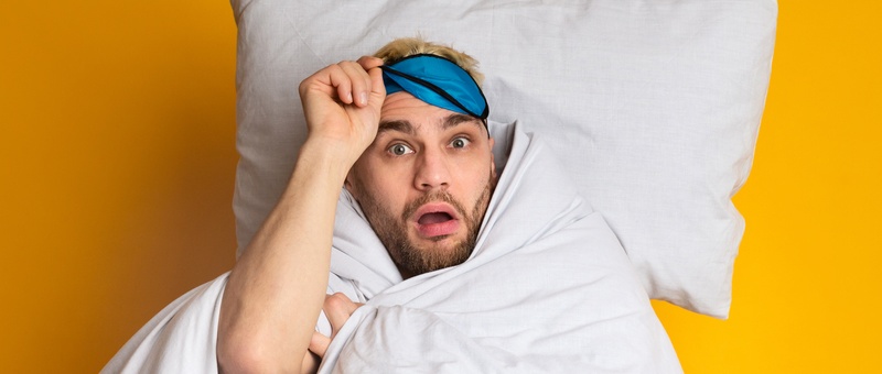 Shocked blonde-haired man in white bedding lifting black and blue sleep mask up from eyeline against a yellow background.