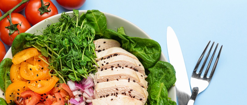Chicken and leafy green salad with onions and tomatoes in bowl beside silver cutlery against a blue background.