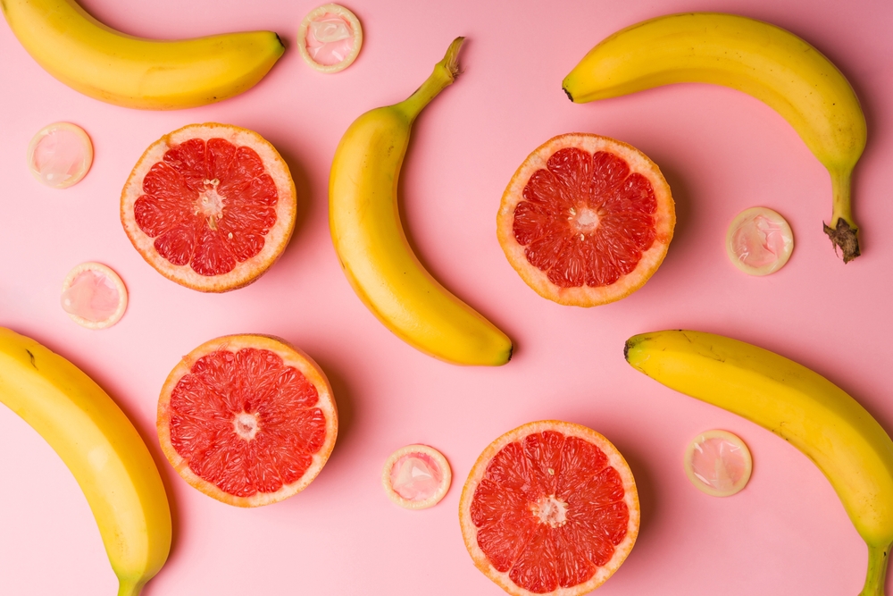 Sliced grapefruits, bananas, and condoms against a pink background.