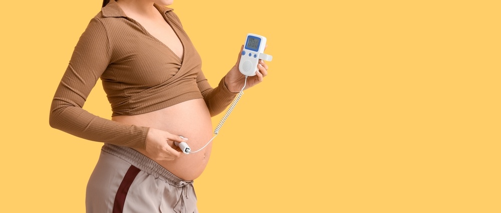 Young pregnant woman with fetal doppler listening to baby's heartbeat on orange background