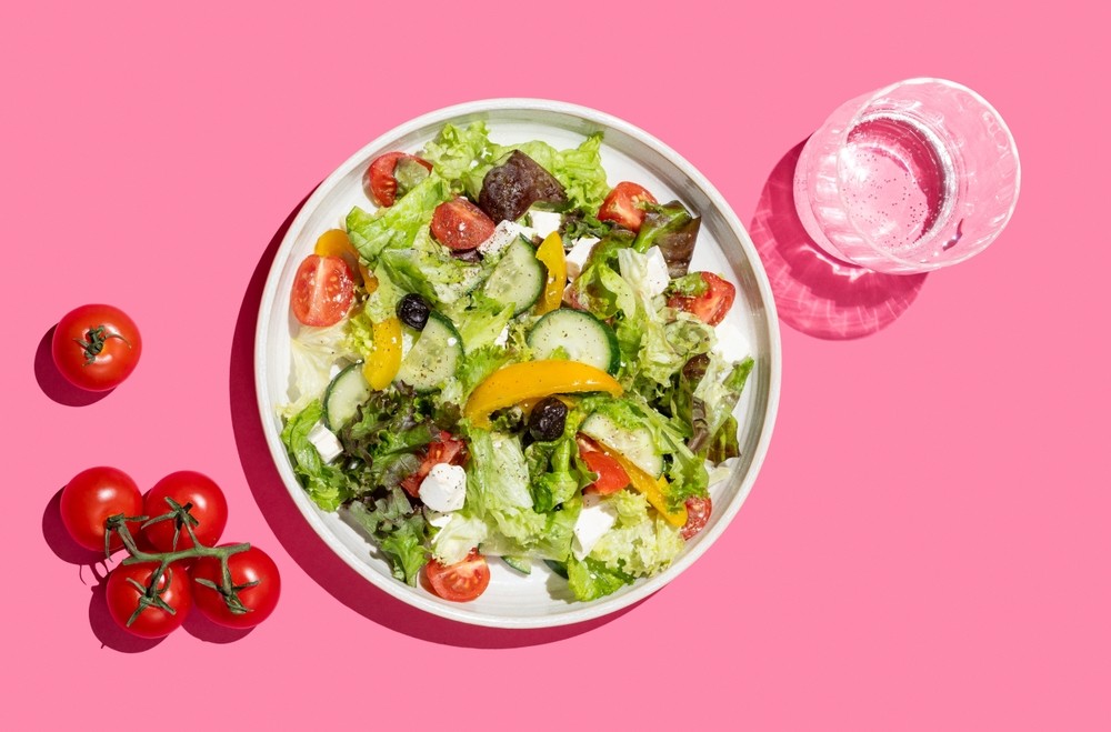 Vegetable salad with lettuce, vegetables, olives and feta cheese on bright pink background with glass of water. Fresh and natural healthy food concept for banner advertising or menu layout. Top view.