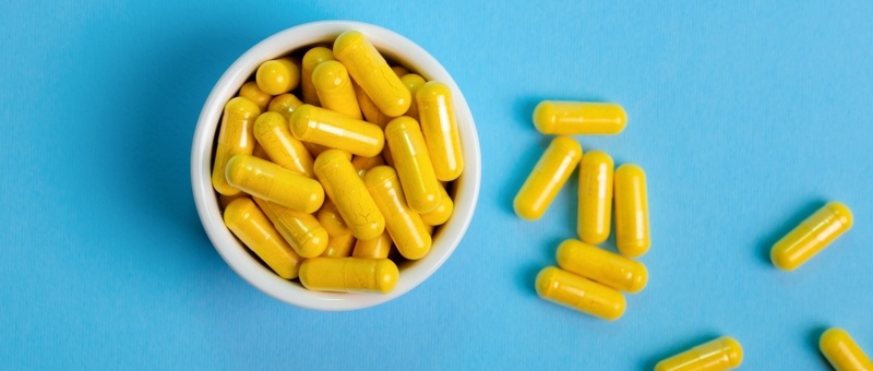 Top view of yellow-coloured supplements in round white ramekin, with some supplements scattered alongside, against a blue background.