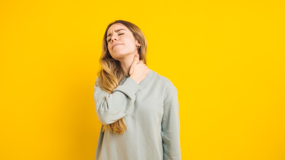 A woman in a light blue sweatshirt holds her neck in pain, eyes closed, against a bright yellow background.