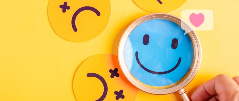 Magnifying glass with red notification-style heart focusing on blue smiley face among yellow sad expressions against yellow background.