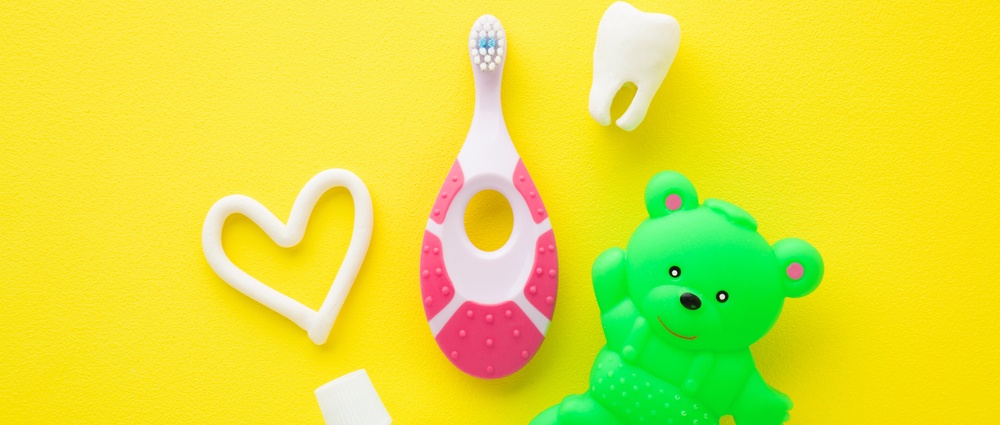 Green rubber teddy bear, white heart shape, tooth, tube of toothpaste, pink toothbrush on bright yellow table background. Things for babies teeth cleaning through fun. Closeup.